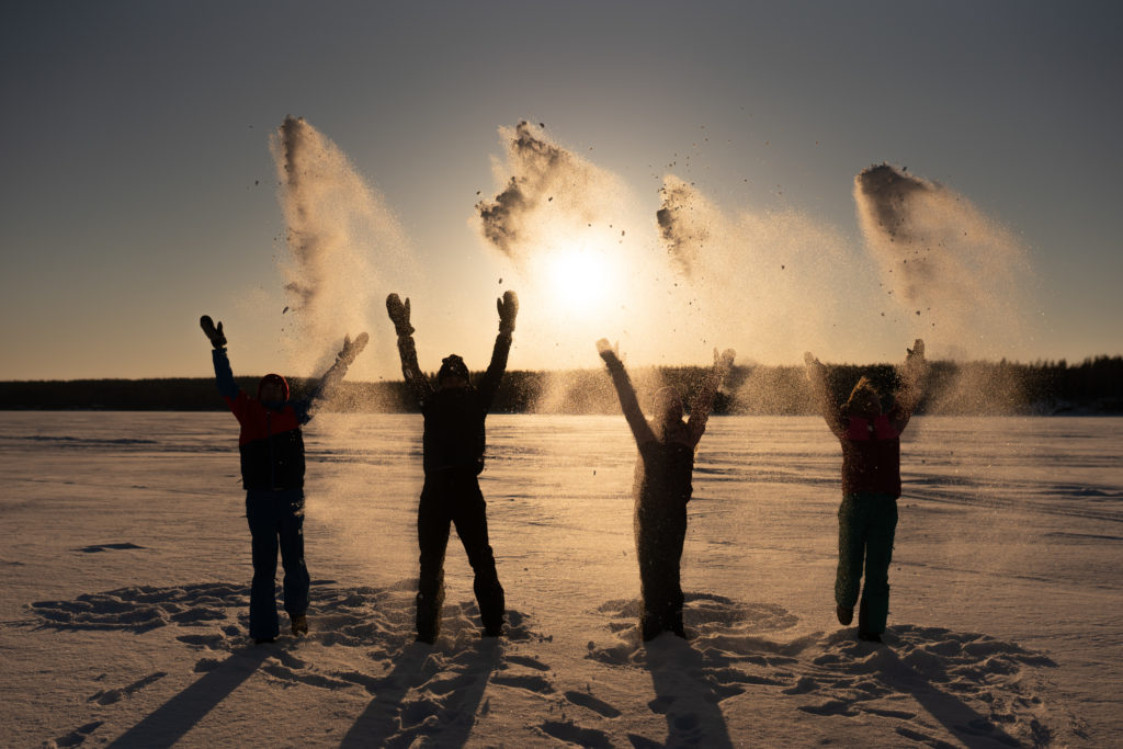 Four young people standing on the snowy lake throwing snowflakes, arms outstretched upwards, winter sunset as the background