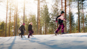 Lapset juoksevat lumikengät jalassa aurinkoisessa ja lumisessa metsässä.