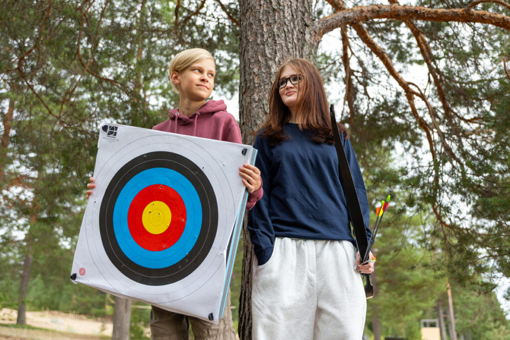 Kaksi nuorta pitelee jousiammunnan maalia kädessään ja nojaa puuhun.